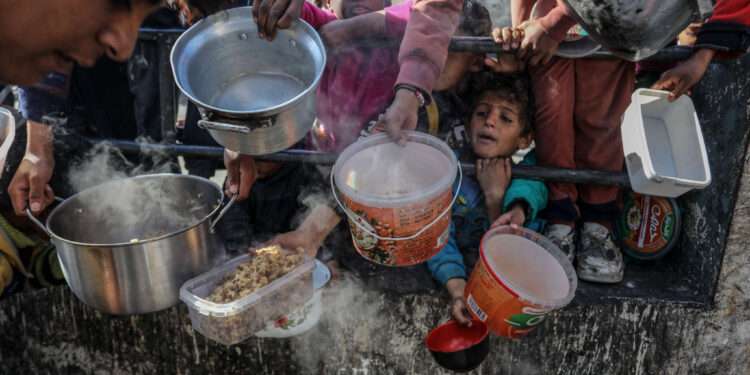 Palestinian children wait in line to receive food prepared by volunteers for Palestinian families displaced to Southern Gaza due to Israeli attacks, between rubble of destroyed buildings in Rafah, Gaza, on February 9, 2024.