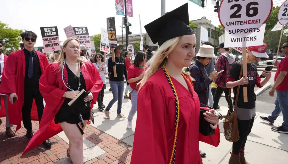 People dressed in commencement gowns, front, walk past protesters supporting the Hollywood writers' strike