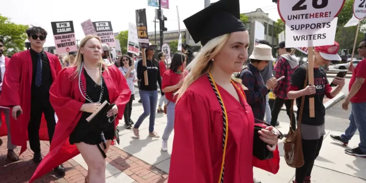 People dressed in commencement gowns, front, walk past protesters supporting the Hollywood writers' strike