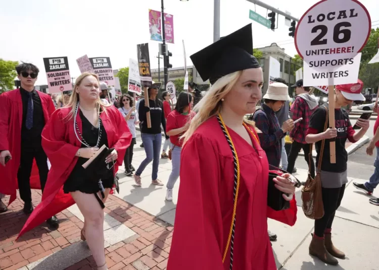 Warner CEO Booed At Boston University As Supporters Of Writers’ Strike Picket Outside 1 People dressed in commencement gowns, front, walk past protesters supporting the Hollywood writers' strike
