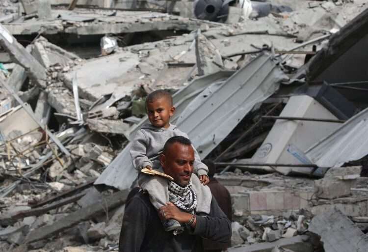 War Wreaks Havoc To Gaza’s Employment Rate 1 A Palestinian man carries his child on his shoulders as he walks past damaged buildings in Khan Younis on April 8, 2024 after Israel pulled its ground forces out of the southern Gaza Strip.